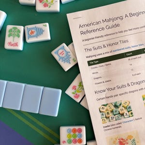 May include: A collection of white Mahjong tiles with colorful designs and text, scattered on a green surface. A reference guide titled "American Mahjong: A Beginning Reference Guide" is also visible, providing information on the game's tiles and rules.