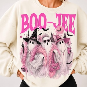 May include: A cream-colored crewneck sweatshirt featuring a graphic print with the text "BOO-JEE" in bright pink. The design depicts three ghosts wearing pink feather boas, hats, and accessories, surrounded by bats, a skull, and a castle.