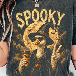 以下が含まれることがあります： 魔女の帽子とハート型のサングラスをかけたガチョウのグラフィックが描かれたチャコールグレーのTシャツ。ガチョウはコーヒーカップを持ち、ピースサインをしています。大きな黄色い月と暗い木々の上に「SPOOKY」という文字が書かれています。