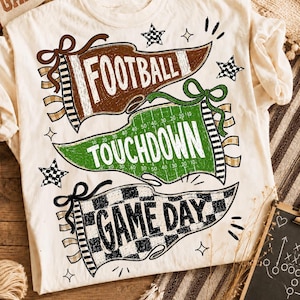Op de afbeelding: Een crèmekleurig T-shirt met drie wimpelvormige afbeeldingen. De bovenste bruine wimpel bevat de tekst FOOTBALL, de middelste groene wimpel met veldlijnen toont TOUCHDOWN en de onderste zwart-wit geblokte wimpel toont de tekst GAME DAY.