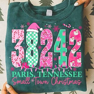 May include: Teal sweatshirt featuring the postal code 38242 in pink and green, with Christmas decorations. The text "Paris, Tennessee Small Town Christmas" is printed below the numbers.