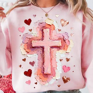 May include: A light pink sweatshirt featuring a large, textured cross design in shades of pink, peach, and cream. Scattered around the cross are various heart shapes in red, pink, and glittery gold. The sweatshirt has long sleeves.