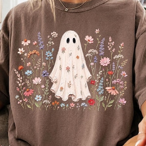 May include: A brown short-sleeve t-shirt featuring a central graphic of a white ghost surrounded by a variety of colorful wildflowers in shades of pink, blue, orange, and purple. The ghost figure is also decorated with small floral patterns.