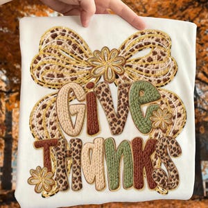 May include: Cream-coloured t-shirt with an embroidered design. The design features the words "GIVE THANKS" in various textures and colours, including brown, green, and tan. A large bow and floral accents complete the festive autumn theme.