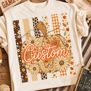 Pode incluir: Uma t-shirt de cor creme com um grande gráfico central de abóbora composto por vários padrões florais, de leopardo e xadrez. A palavra Custom está impressa numa fonte cursiva laranja arrojada no centro da abóbora, sobre riscas verticais.