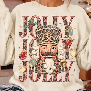 May include: A cream-coloured crewneck sweatshirt featuring a large graphic of a nutcracker wearing a leopard print uniform. The text JOLLY is printed in a matching leopard pattern behind the nutcracker, accented by red and green Christmas lights.