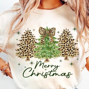 May include: White t-shirt with a graphic of three Christmas trees decorated with leopard print and lights. The trees are topped with a bow and the text "Merry Christmas" is printed below.