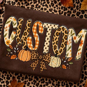 以下が含まれることがあります： 大きなブロック体で「CUSTOM」と書かれたダークブラウンのTシャツ。各文字には花柄、ヒョウ柄、オレンジ色のチェック柄などのユニークなパターンが施されています。文字の下には2つのカボチャとヒョウ柄のリボン、秋の葉が描かれています。