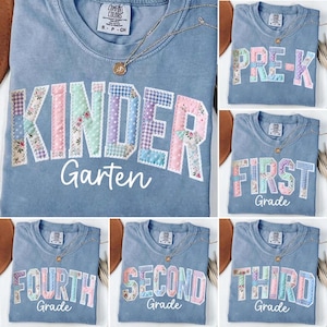 May include: A light blue Comfort Colors t-shirt with "KINDER Garten" in patchwork letters. The shirt has a gold necklace. Other shirts show "PRE-K", "FIRST Grade", "FOURTH Grade", "SECOND Grade", and "THIRD Grade".