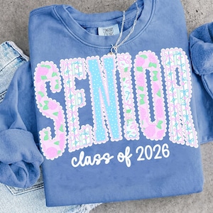 May include: A blue sweatshirt with the word "SENIOR" in a floral design with a light blue "N" and the words "class of 2026" in white. The sweatshirt has a Comfort Colors tag.