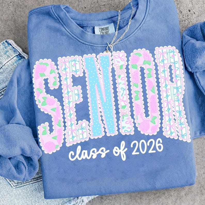 May include: A blue sweatshirt with the word "SENIOR" in a floral design with a light blue "N" and the words "class of 2026" in white. The sweatshirt has a Comfort Colors tag.