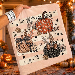 May include: A light pink square fabric item featuring a printed design of three pumpkins. One pumpkin is orange with a white floral pattern, one is tan with a leopard print and black bow, and one is black with a white floral pattern and a bow.