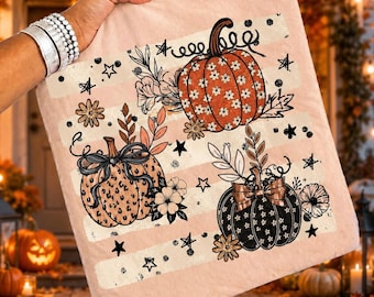 Puede incluir: Un artículo de tela cuadrado de color rosa claro con un diseño estampado de tres calabazas. Una calabaza es naranja con un patrón floral blanco, una es beige con estampado de leopardo y lazo negro, y una es negra con patrón floral blanco y lazo.