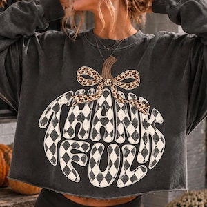 May include: A charcoal gray long-sleeve sweatshirt with a raw-edge hem. The front features a large pumpkin graphic with a black and white checkered pattern, topped with a leopard print bow and the word THANKFUL written in bold white letters.