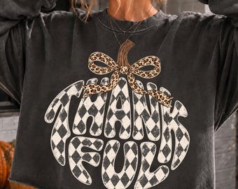 May include: A charcoal gray long-sleeve sweatshirt with a raw-edge hem. The front features a large pumpkin graphic with a black and white checkered pattern, topped with a leopard print bow and the word THANKFUL written in bold white letters.