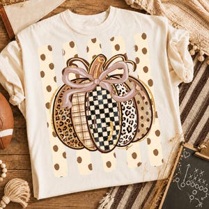 K&ouml;nnte beinhalten: Ein cremefarbenes T-Shirt mit einer zentralen Grafik eines K&uuml;rbisses mit einer hellvioletten Schleife. Der K&uuml;rbis ist in vertikale Segmente mit Leopardenmuster, Karomuster und Schachbrettmuster unterteilt, vor einem Hintergrund aus beigen Punkten.