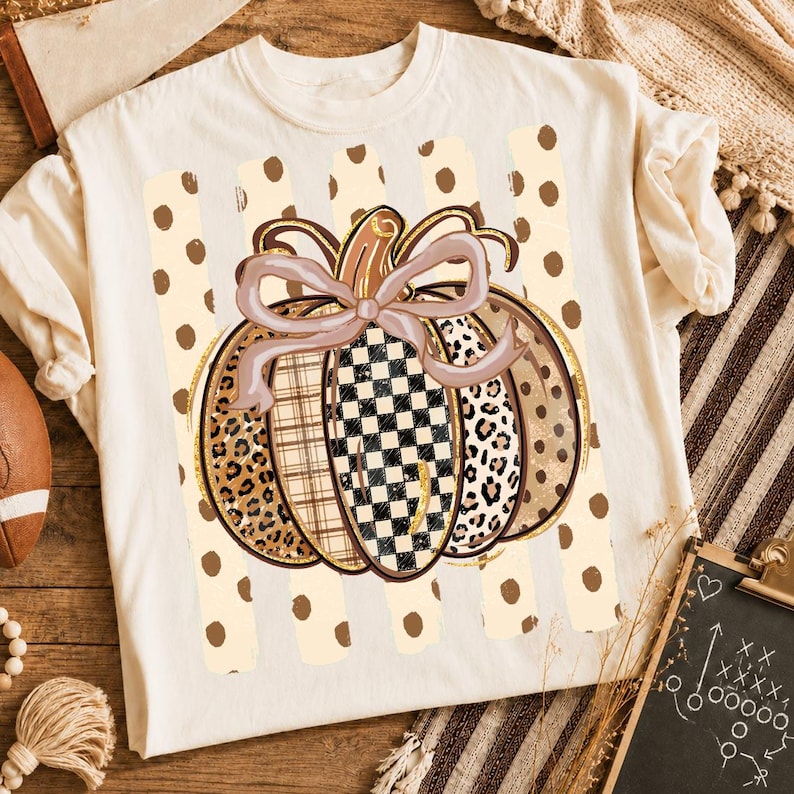 K&ouml;nnte beinhalten: Ein cremefarbenes T-Shirt mit einer zentralen Grafik eines K&uuml;rbisses mit einer hellvioletten Schleife. Der K&uuml;rbis ist in vertikale Segmente mit Leopardenmuster, Karomuster und Schachbrettmuster unterteilt, vor einem Hintergrund aus beigen Punkten.
