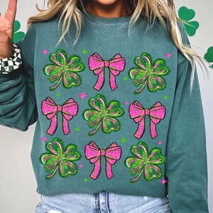 May include: Teal green sweatshirt featuring a pattern of pink bows and green shamrocks. The design includes glitter accents and is likely themed for St. Patrick's Day. The sweatshirt is made of a soft material.