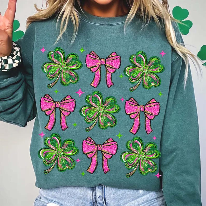 May include: Teal green sweatshirt featuring a pattern of pink bows and green shamrocks. The design includes glitter accents and is likely themed for St. Patrick's Day. The sweatshirt is made of a soft material.