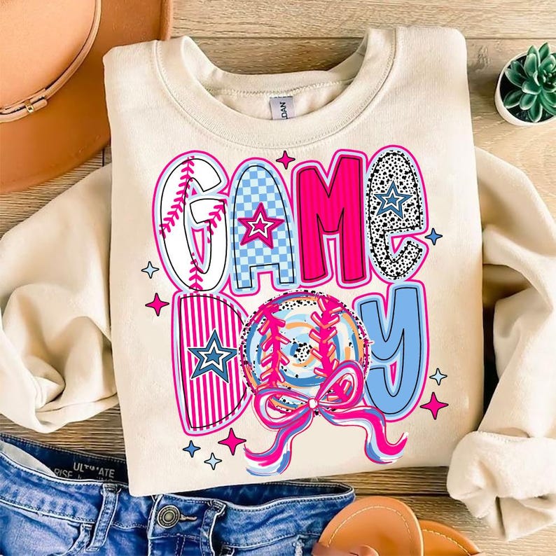 May include: A cream-colored sweatshirt with the text "GAME DAY" in pink, blue, and white. The text is decorated with stars, baseball stitching, and a baseball graphic.