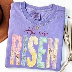 May include: A lavender Comfort Colors t-shirt with the words "He is Risen" in a patchwork design. The letters are made of various patterned fabrics, including gingham and floral prints. A silver necklace with a small pendant rests on the shirt.