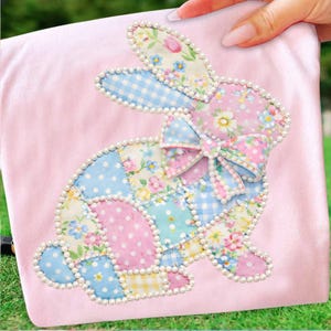 May include: A pink fabric with a patchwork bunny design. The bunny is made of various floral and polka dot fabrics, outlined with faux pearls. A bow adorns the bunny's neck. The design is held by a hand.