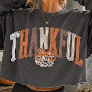 May include: A charcoal gray crewneck sweatshirt featuring the word THANKFUL in a stylized, multi-line font. The letter K is replaced by a graphic of a white pumpkin with an orange bow. The text uses a mix of white and orange line-art lettering.