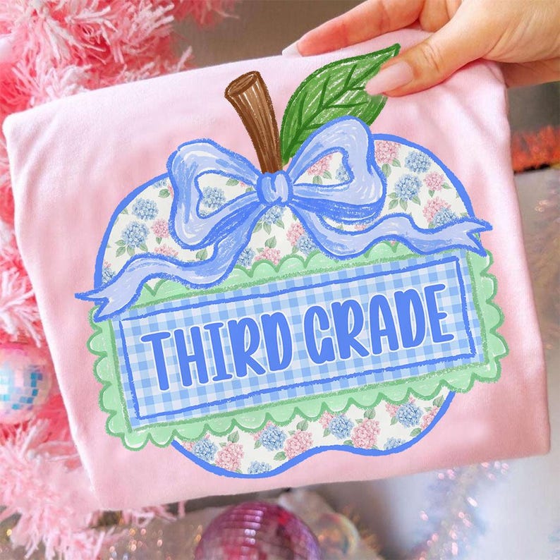 Op de afbeelding: Een lichtroze t-shirt met een afbeelding van een appel met bloemenpatroon, een bruine steel en een groen blad. Een blauw lint is rond de steel gestrikt. Een blauw-wit geblokte banner in het midden bevat de tekst THIRD GRADE in blauwe letters.