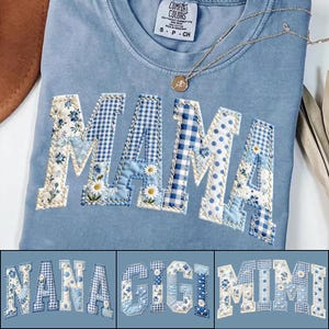 Op de afbeelding: Lichtblauw Comfort Colors T-shirt met het woord "MAMA" in patchworkstof. De letters hebben bloemen-, ruit- en stippatronen in blauw- en wittinten. Een gouden ketting met een hanger is over het shirt gedrapeerd. Extra shirts met "NANA", "GIGI" en "MIMI" worden getoond.