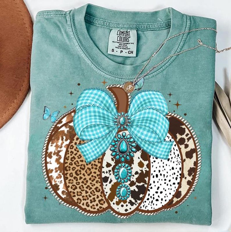 May include: A teal Comfort Colors t-shirt featuring a pumpkin design. The pumpkin has a brown stem, a turquoise gingham bow, and a pattern of leopard print, cowhide, and white spots. A turquoise necklace and a butterfly are also visible.