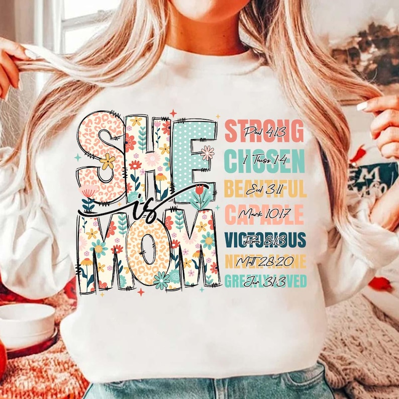 K&ouml;nnte beinhalten: Wei&szlig;er Pullover mit einem Blumendesign, auf dem "She is Mom" steht, darunter aufgelistete Bibelverse. Die Verse sind: Phil 4:13, 1 Thess 1:4,  Ed 3:11, Mark 10:17,  Ps 28:13, Mt 28:20,  Jn 3:13.