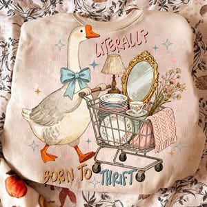 May include: A light pink sweatshirt with a whimsical illustration of a goose wearing a blue bow tie, pushing a shopping cart filled with vintage items. Text reads "Literally Born to Thrift."