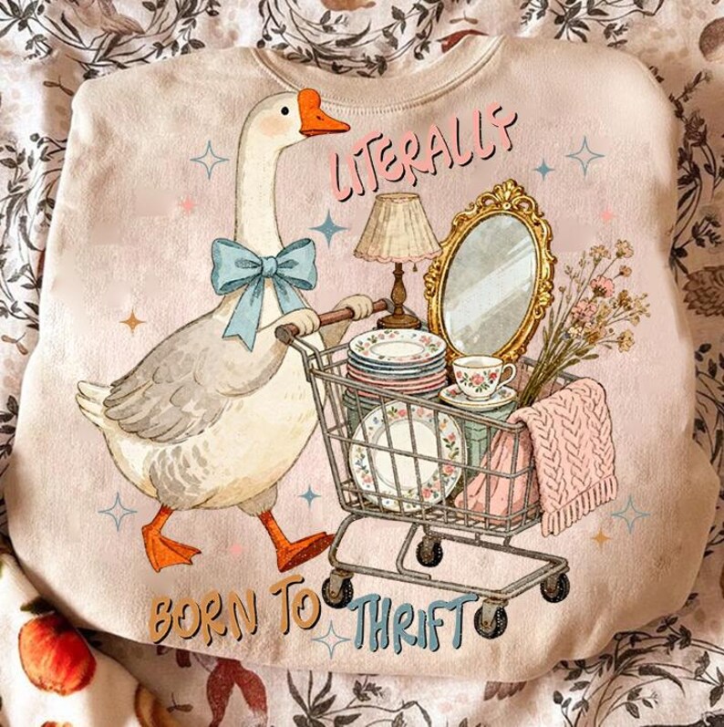 May include: A light pink sweatshirt with a whimsical illustration of a goose wearing a blue bow tie, pushing a shopping cart filled with vintage items. Text reads "Literally Born to Thrift."