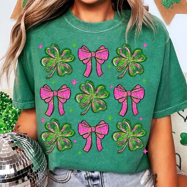 May include: Green t-shirt featuring a pattern of pink bows and green shamrocks. The shamrocks have a green glitter effect, and the bows have a pink glitter effect. The shirt is a crew neck style.