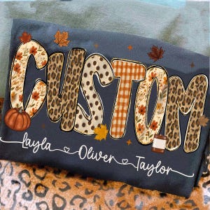 May include: A dark gray t-shirt featuring the word CUSTOM in large, patterned letters with floral, leopard print, polka dot, and orange gingham designs. An orange pumpkin, brown leaves, and the names Layla, Oliver, and Taylor are printed below.