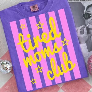 Puede incluir: Una camiseta morada con rayas rosas verticales y texto amarillo que dice "tired moms club". Estrellas amarillas est&aacute;n dispersas alrededor del texto. La camiseta tiene una etiqueta Comfort Colors.