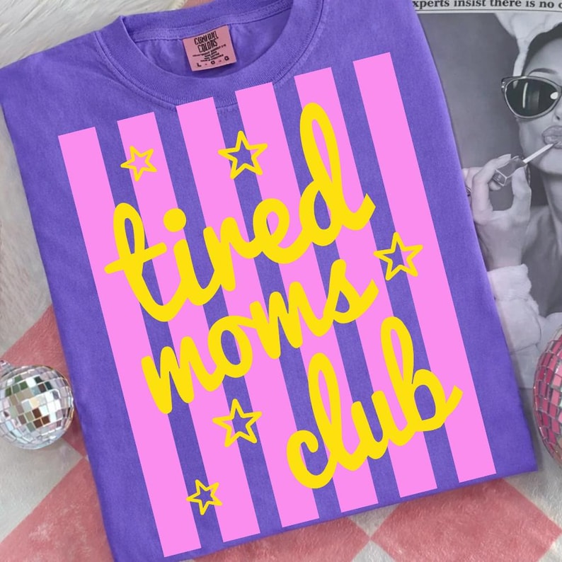 Puede incluir: Una camiseta morada con rayas rosas verticales y texto amarillo que dice "tired moms club". Estrellas amarillas est&aacute;n dispersas alrededor del texto. La camiseta tiene una etiqueta Comfort Colors.