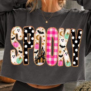 May include: A charcoal gray long-sleeve sweatshirt featuring the word SPOOKY in large letters. Each letter has a unique pattern including black and white polka dots, pink and white gingham, and illustrations of ghosts, bats, and pumpkins.