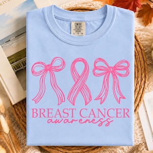 May include: A light blue t-shirt featuring a graphic print of two pink line-art bows flanking a pink awareness ribbon. Below the graphic, the text reads BREAST CANCER awareness in pink lettering. The shirt has a Comfort Colors brand neck label.