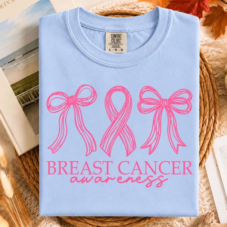 May include: A light blue t-shirt featuring a graphic print of two pink line-art bows flanking a pink awareness ribbon. Below the graphic, the text reads BREAST CANCER awareness in pink lettering. The shirt has a Comfort Colors brand neck label.
