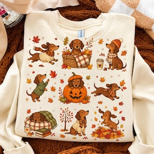 Könnte beinhalten: Ein cremefarbenes Sweatshirt mit Rundhalsausschnitt und einem wiederkehrenden Muster aus braunen Dackeln bei verschiedenen herbstlichen Aktivitäten, darunter das Tragen von Pullovern, Sitzen in einem Kürbis, Spielen im Laub und Ausruhen in einem karierten Hundebett.
