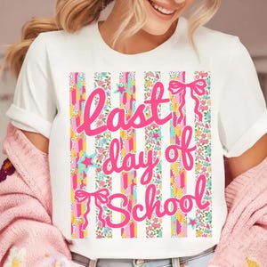 Puede incluir: Camiseta blanca con un dise&ntilde;o de rayas coloridas con estampados florales, estrellas y lazos. Las palabras "Last day of School" est&aacute;n escritas en rosa en la parte delantera. Se lleva una rebeca rosa sobre la camiseta.