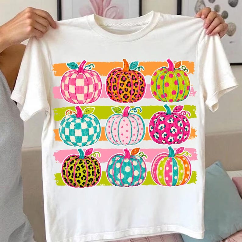 May include: A white short-sleeve t-shirt featuring a graphic print of nine pumpkins arranged in a three-by-three grid. The pumpkins display various patterns including checkered, leopard print, and polka dots in vibrant pink, orange, teal, and green.