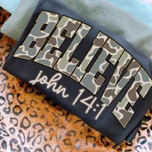 May include: A folded black t-shirt with the word "BELIEVE" in large, camouflage-patterned letters. The text "john 14:1" is printed below in a cursive font. The shirt is layered with other folded shirts on a leopard print surface.