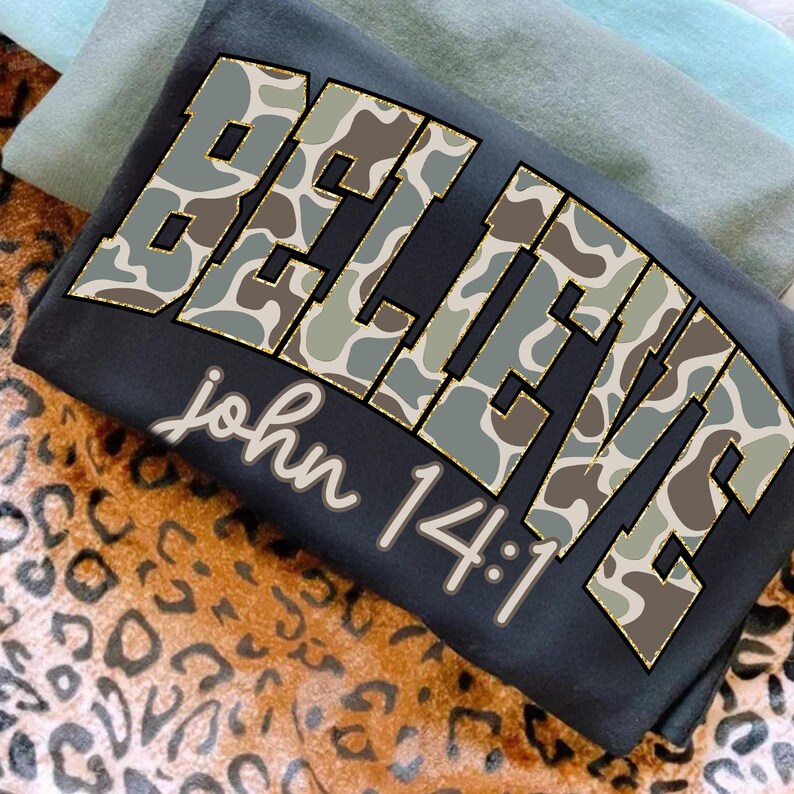 May include: A folded black t-shirt with the word "BELIEVE" in large, camouflage-patterned letters. The text "john 14:1" is printed below in a cursive font. The shirt is layered with other folded shirts on a leopard print surface.