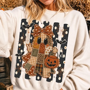 May include: A cream-colored sweatshirt featuring a central graphic of a patchwork ghost wearing a large bow. The ghost is holding a jack-o'-lantern bucket and making a peace sign. The background includes black vertical stripes with white polka dots, spiderwebs, and small black bats.