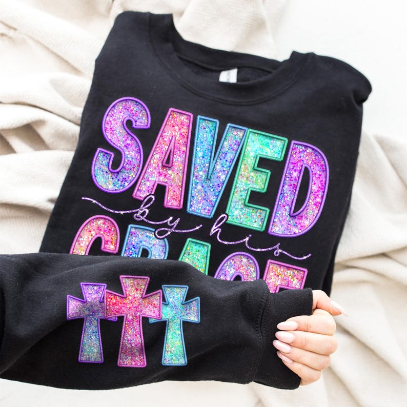 May include: Black sweatshirt with the words "SAVED by his GRACE" in colorful glitter lettering. The sleeve displays three glitter cross designs in purple, pink, and blue. The sweatshirt is displayed on a neutral background.