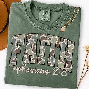 May include: A sage green t-shirt with the word "FAITH" in a camouflage pattern with gold glitter trim. The text "ephesians 2:8" is below the word "FAITH". A gold necklace with a round pendant is draped on the shirt.