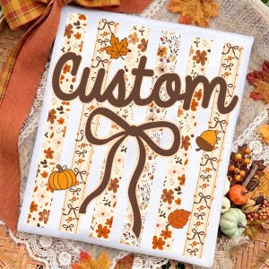Op de afbeelding: Een wit t-shirt met verticale strepen met herfstachtige bloemen- en bladpatronen in oranje, bruin en cr&egrave;me. Het woord Custom is in bruin cursief gedrukt over het midden, geplaatst over een grote bruine afbeelding van een gestrikte strik.