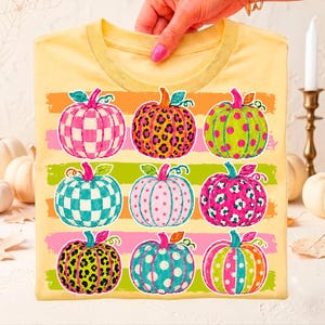 May include: A pale yellow t-shirt featuring a graphic print of nine pumpkins arranged in three rows. Each pumpkin displays a unique pattern, including checkered, leopard print, and polka dot designs in vibrant shades of pink, green, and orange.